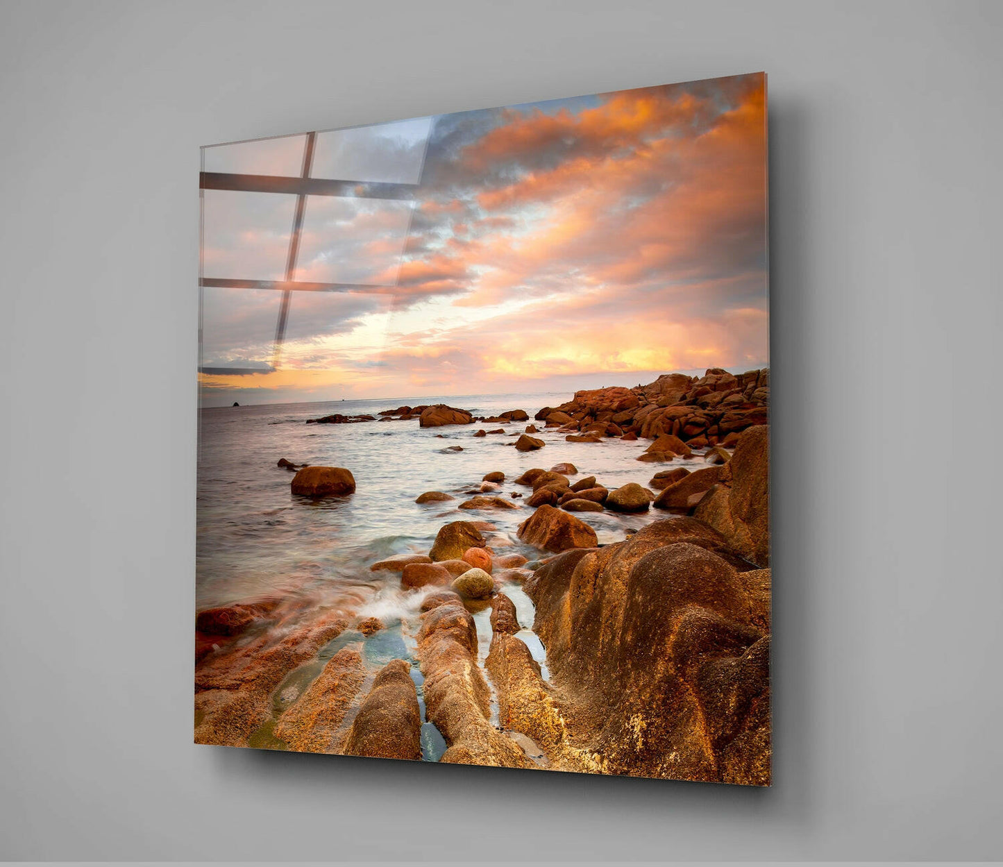 Glass wall art displaying a rocky coastal sunset with golden-orange sky and boulder-strewn beach, mounted on gray wall.
