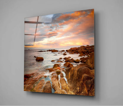 Glass wall art displaying a rocky coastal sunset with golden-orange sky and boulder-strewn beach, mounted on gray wall.