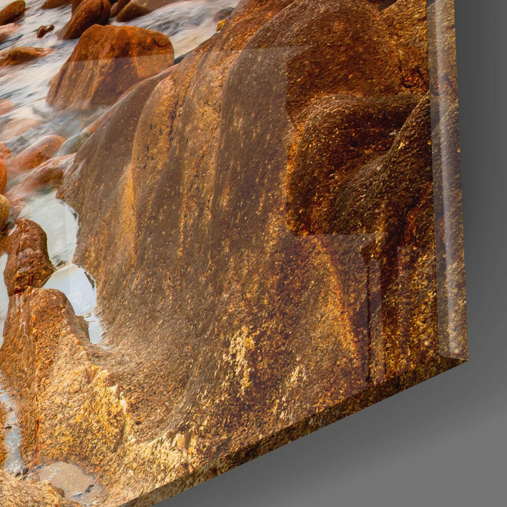 Close-up of glass wall art edge showing rust-colored rocks and flowing water with golden-brown tones and textured surface detail.