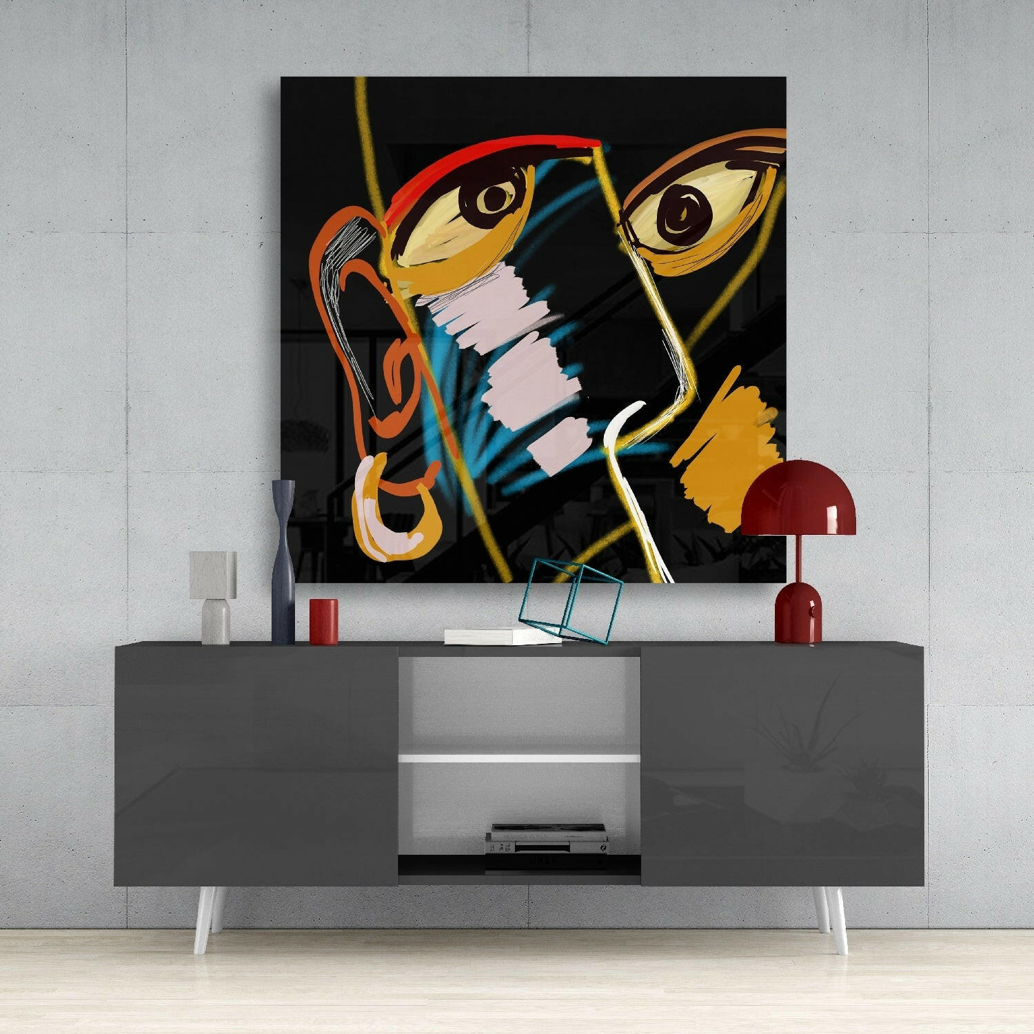 Bold abstract artwork with stylized faces in red, orange, yellow, and blue on black glass canvas displayed above gray media console with red lamp.