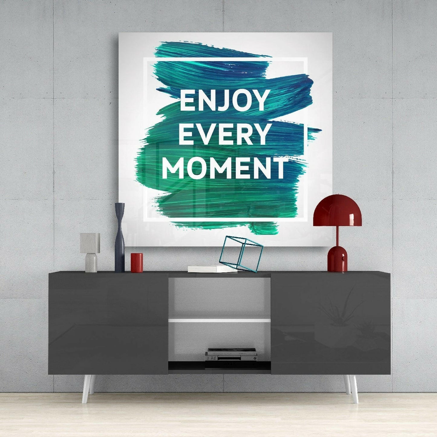Glass wall art with teal brush strokes displaying "ENJOY EVERY MOMENT" text mounted above dark console table.