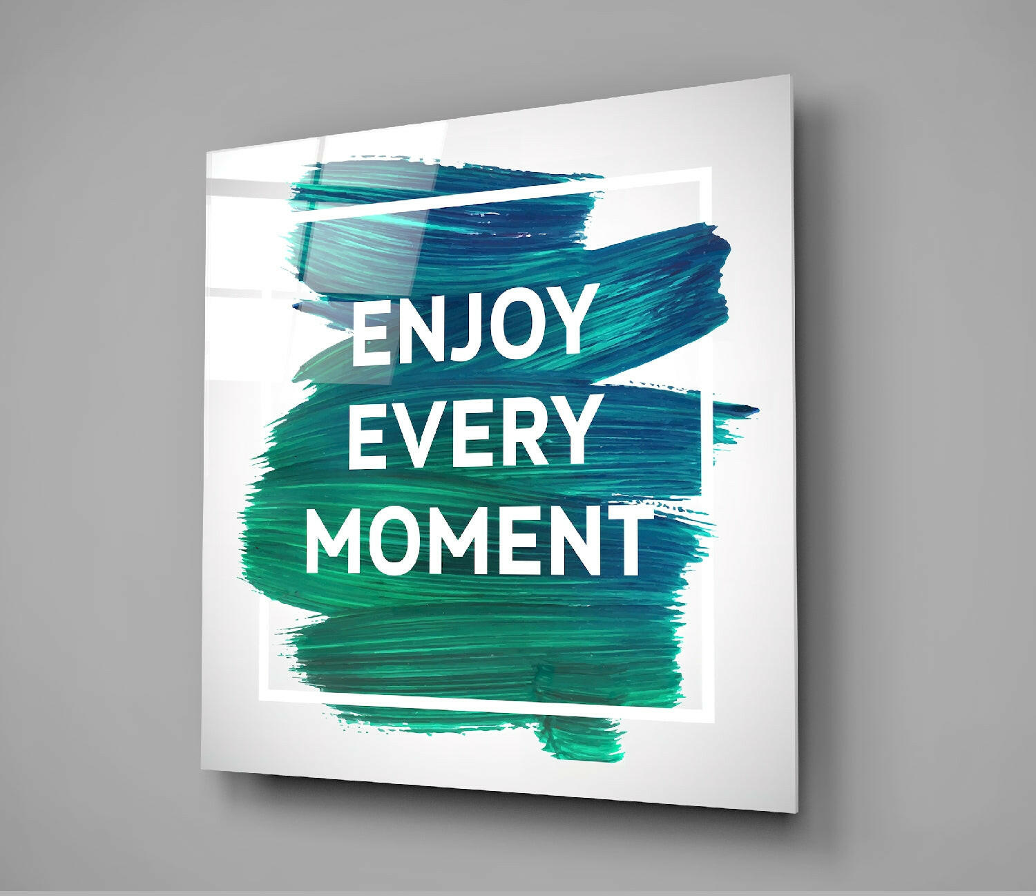 Glass wall art panel displaying white text "ENJOY EVERY MOMENT" overlaid on teal and turquoise brush strokes, angled perspective view.