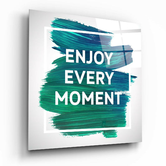 Glass wall art panel displaying "ENJOY EVERY MOMENT" in white text over teal and green brush strokes, angled perspective view.