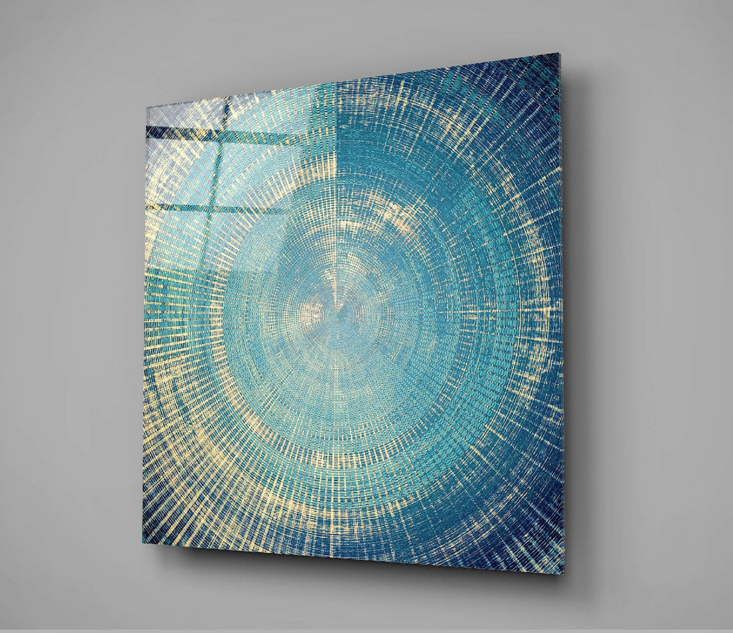 Square glass wall art with concentric circular pattern in teal, navy, and gold tones, mounted at angle on gray wall.