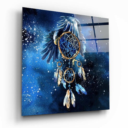 Angled view of glass wall art displaying an eagle dreamcatcher with blue feathers, golden rings, and white feathers against a deep blue splattered background.