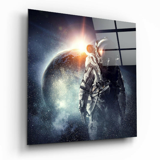 Angled view of glass wall art showing astronaut in spacesuit facing illuminated planet with golden sunrise and starfield, mounted on white wall with black grid pattern.