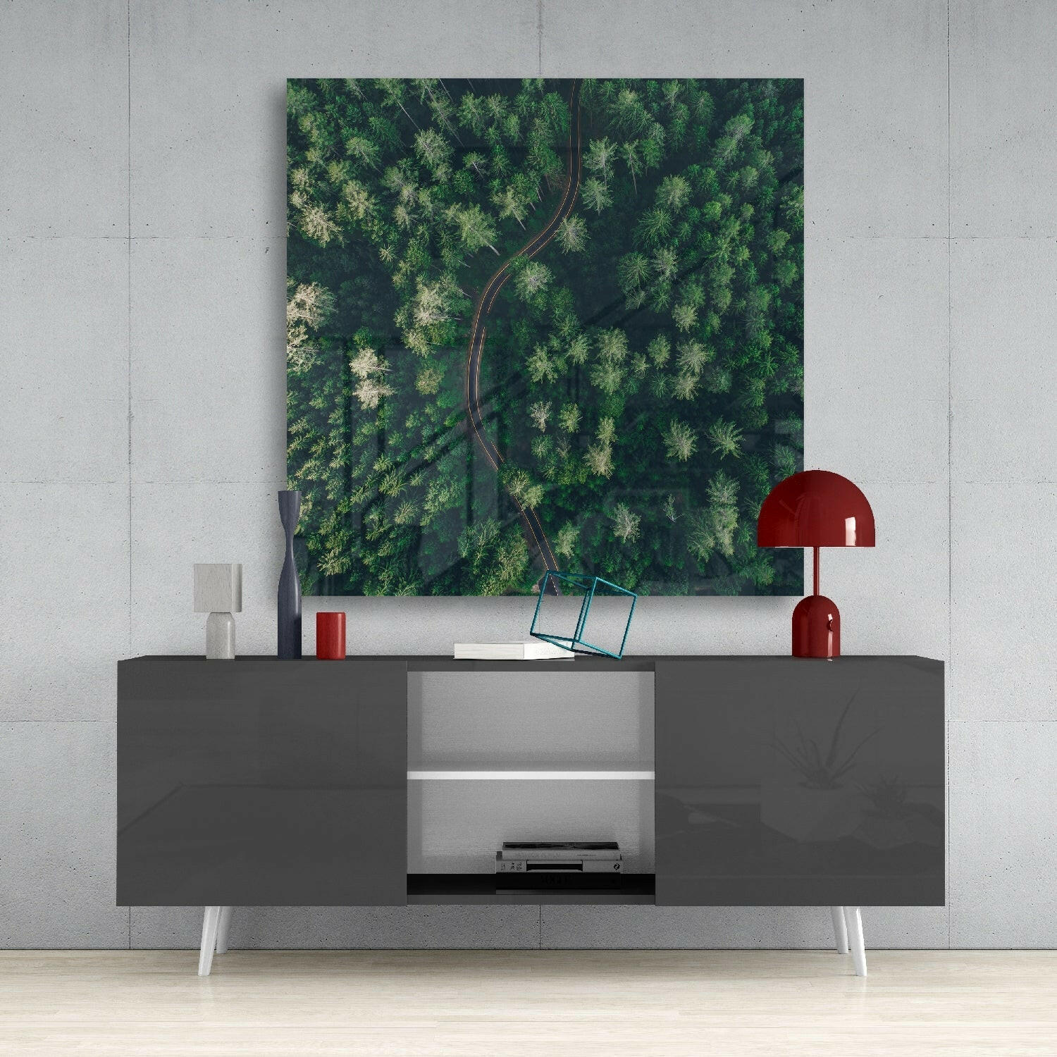 Aerial forest road glass art displayed above gray sideboard with red lamp and accessories in modern interior setting.