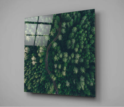 Angled view of glass wall art displaying aerial forest landscape with winding red road and dark grid overlay in upper left corner.