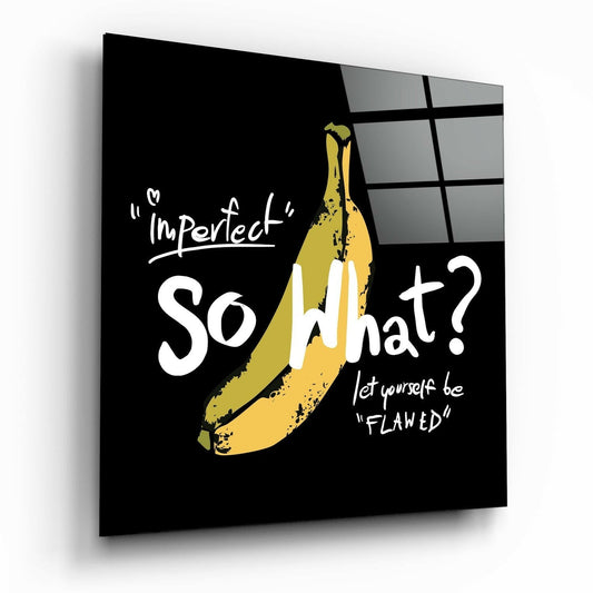 Angled perspective of black glass wall art with yellow banana illustration and white text reading "Imperfect So What? let yourself be Flawed" against gridded window background.