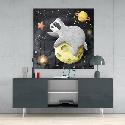 Glass wall art depicting a white sloth relaxing on a yellow moon surrounded by stars and planets against a dark starry background, displayed above a dark gray media console.