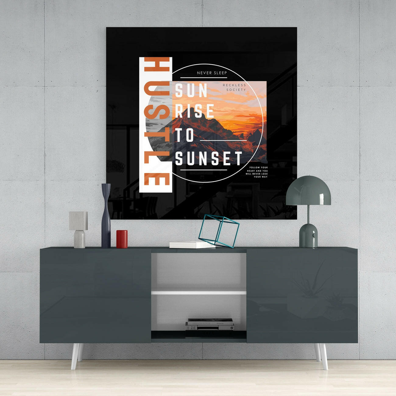 Modern living room setup featuring a dark gray media console with white legs displaying a large black glass wall art print with orange sunset mountain imagery and white text reading "Sunrise to Sunset.