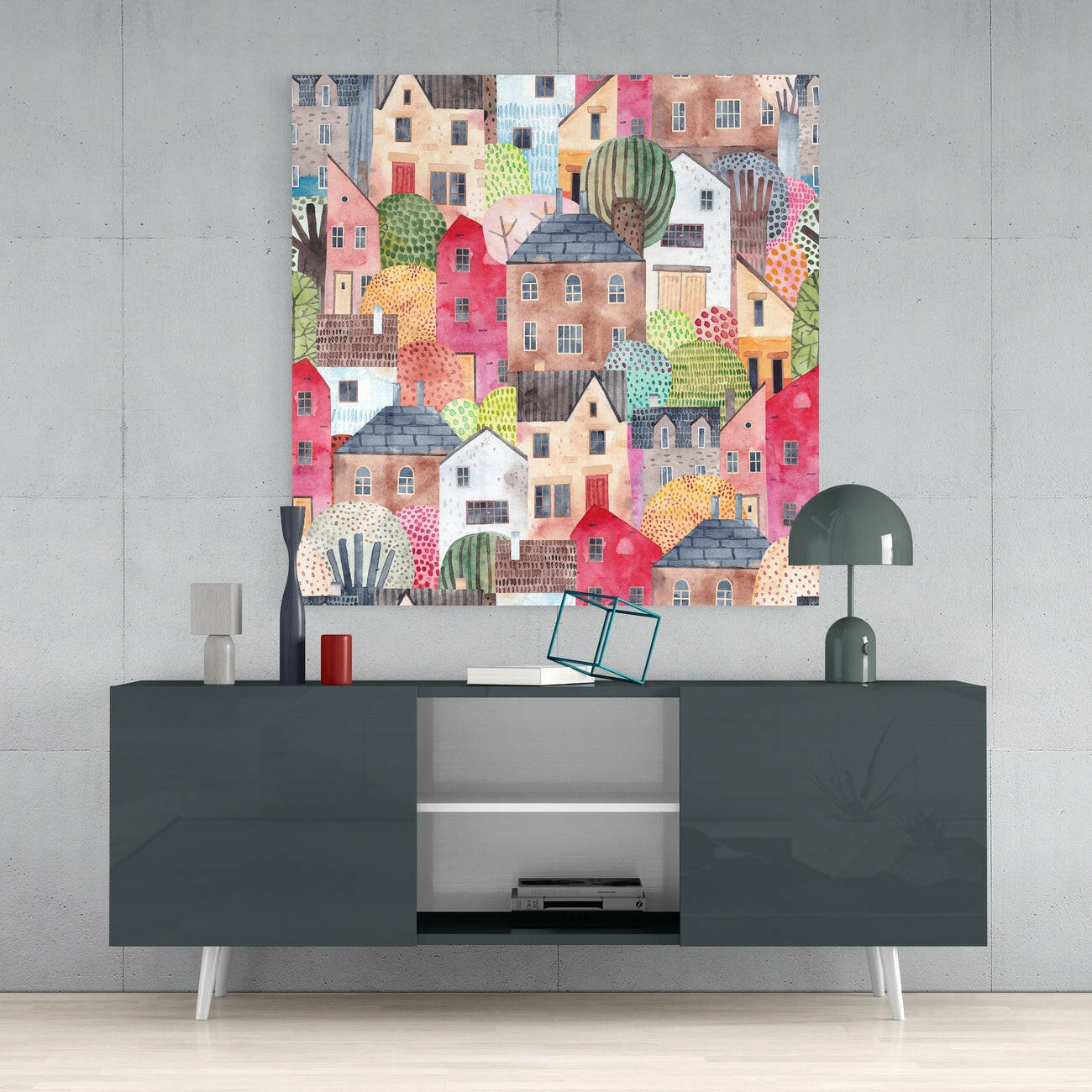 Colorful glass wall art featuring densely packed illustrated houses in pink, red, orange, and earth tones with green cacti, displayed above a dark gray sideboard in a modern interior.