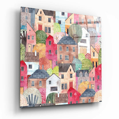 Angled view of a square glass wall art panel featuring a whimsical collage of colorful houses and cacti in warm earth tones and vibrant pinks.