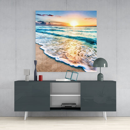 Glass wall art depicting a beach sunset with golden waves and orange sky displayed above a dark gray media console in a modern interior setting.