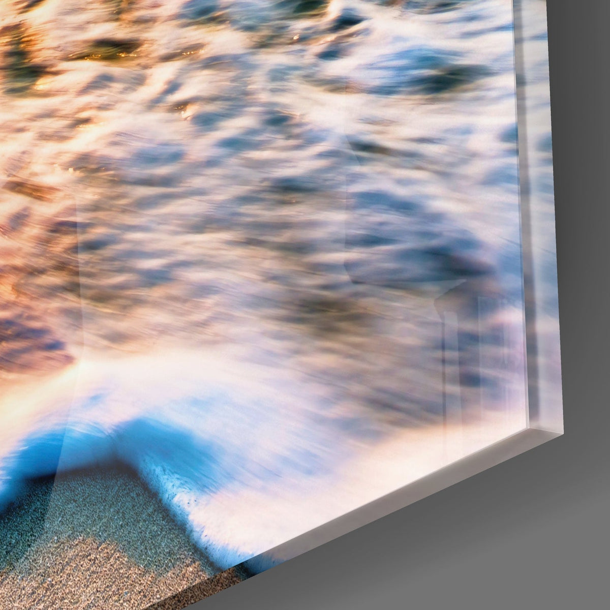 Glass wall art panel displaying ocean waves with golden sunlight, blue water, and white foam on sandy beach, angled view showing edge.