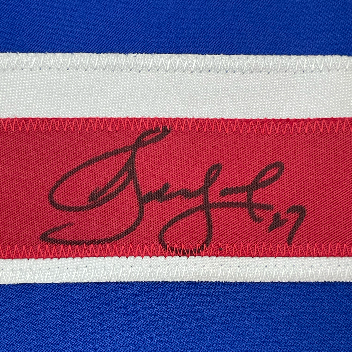 Close-up of autographed red hockey jersey stripe showing handwritten signature in black ink on fabric.