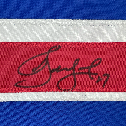 Close-up of autographed red hockey jersey stripe showing handwritten signature in black ink on fabric.