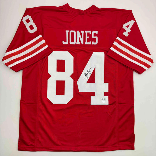 Red San Francisco 49ers football jersey number 84 with "JONES" nameplate, displayed flat showing back view with white striping on sleeves and autographed signature.