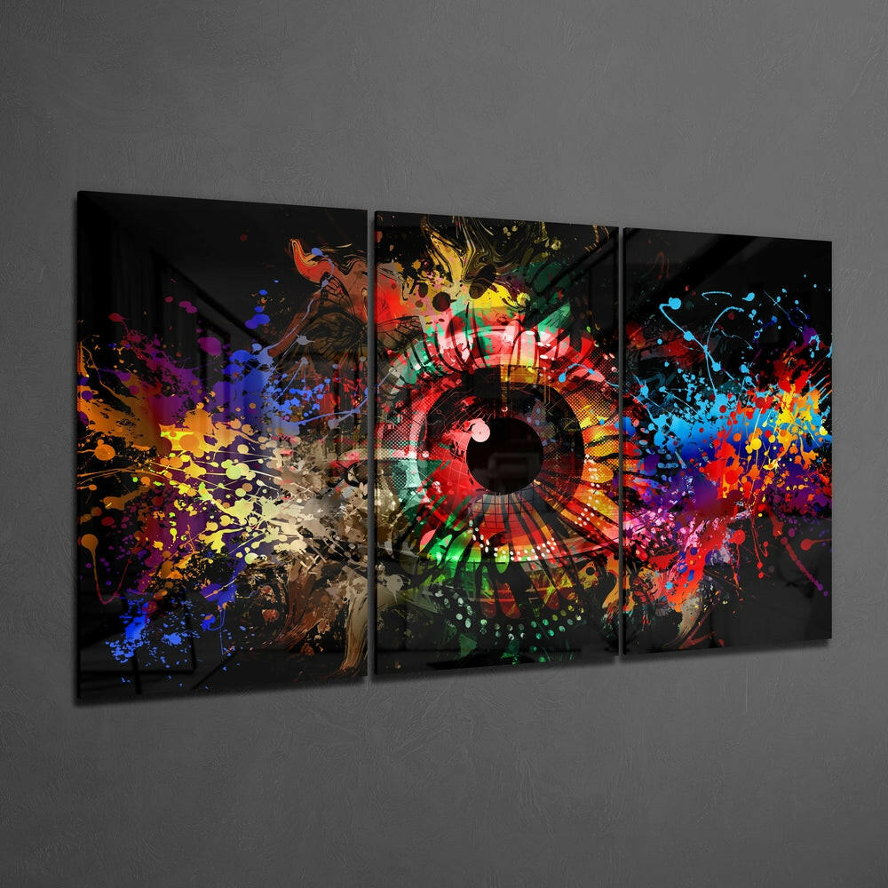Three-panel glass wall art displaying a colorful abstract eye design with vibrant paint splatters in red, blue, yellow, and green on black background, mounted on gray wall.