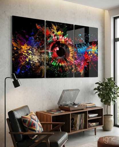 Three-panel colorful glass wall art with abstract eye design and vibrant paint splatters displayed above mid-century furniture in modern interior.