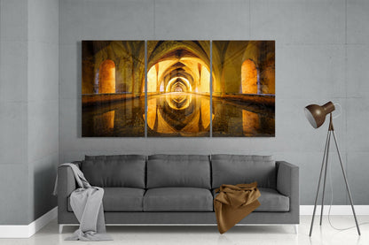 Three-panel glass wall art depicting a symmetrical golden-lit arched stone corridor with water reflection, displayed above a gray sofa in a modern living room.