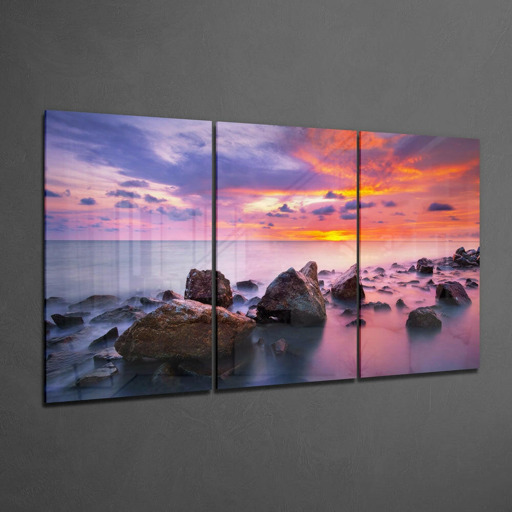 Three-panel glass wall art displaying a rocky coastline at sunset with pink and orange sky, mounted on gray wall.