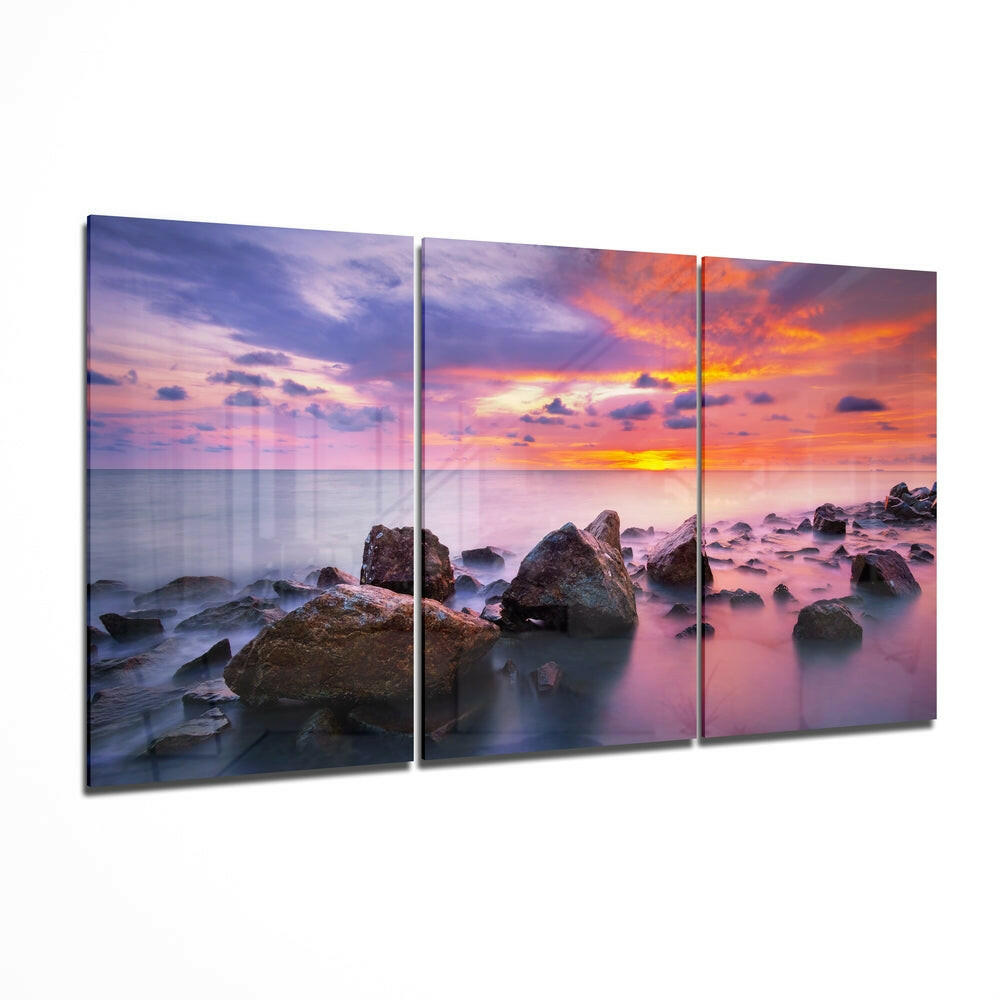 Three-panel glass wall art displaying a rocky coastline at sunset with purple, orange, and pink sky reflections in calm water.