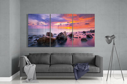 Three-panel glass wall art displaying a vibrant sunset over rocky coastline, installed above gray sofa in minimalist interior.