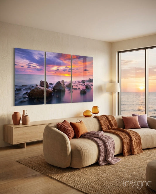 Three-panel glass sunset wall art above wooden credenza in modern coastal living room with ocean view windows.