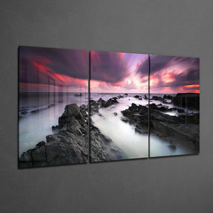 Three-panel glass wall art displaying a rocky coastline at sunset with pink and purple sky, soft misty water, and dark stone formations.