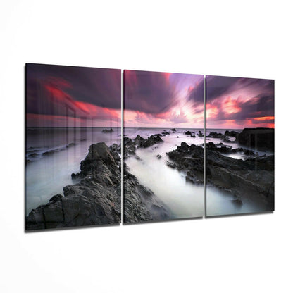Three-panel glass wall art displaying a rocky coastline at sunset with pink and purple sky, misty waves, and dark stones.