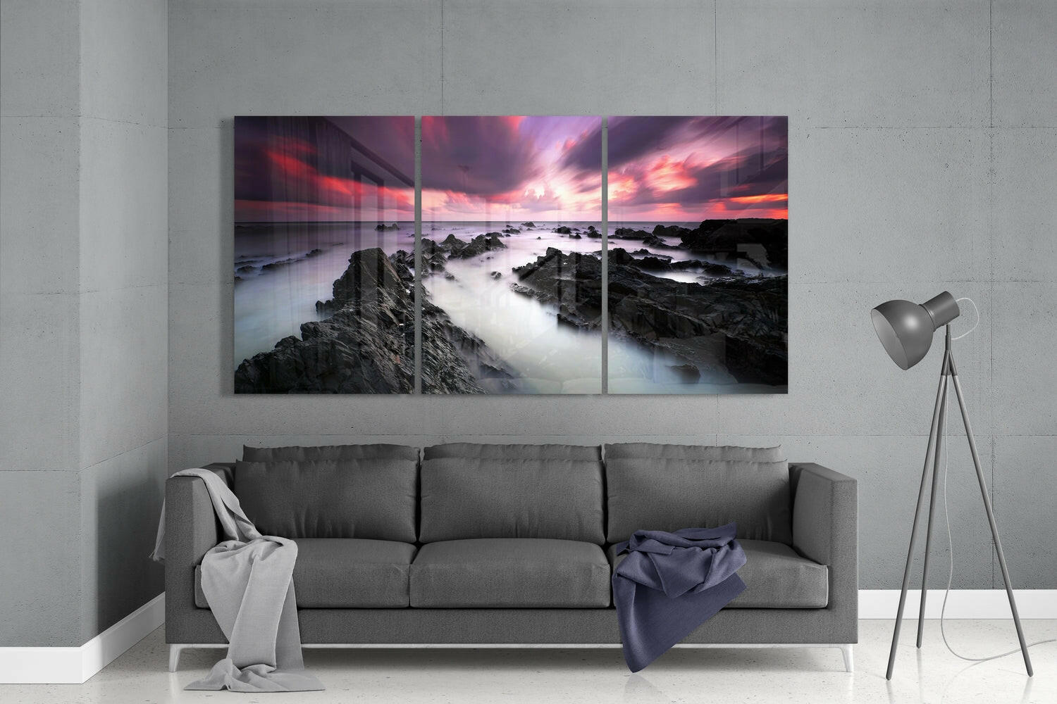 Three-panel glass wall art above gray sofa displaying rocky coastline at sunset with pink and orange sky.