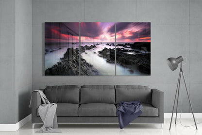Three-panel glass wall art above gray sofa displaying rocky coastline at sunset with pink and orange sky.