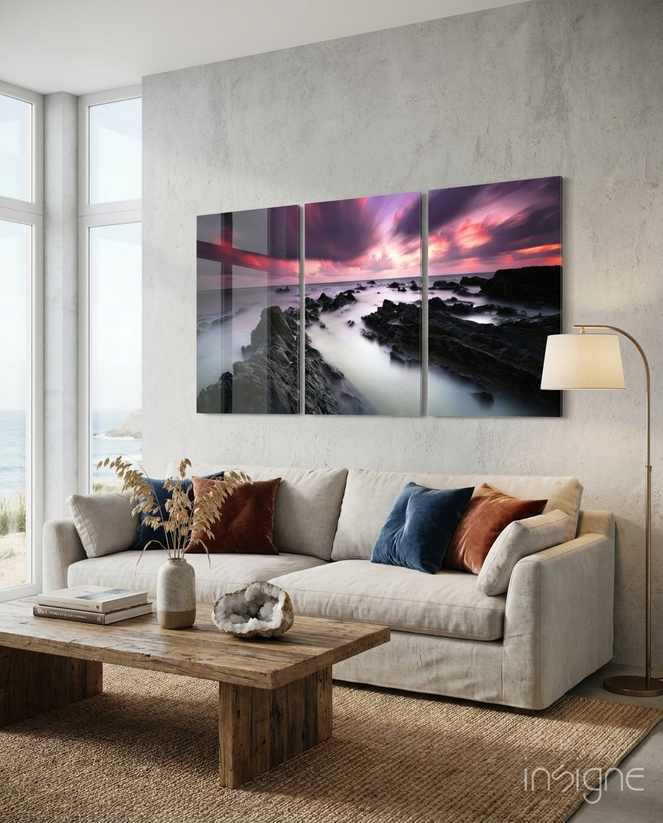 Three-panel glass wall art above beige sofa depicting rocky coastline with pink sunset sky and misty waves.
