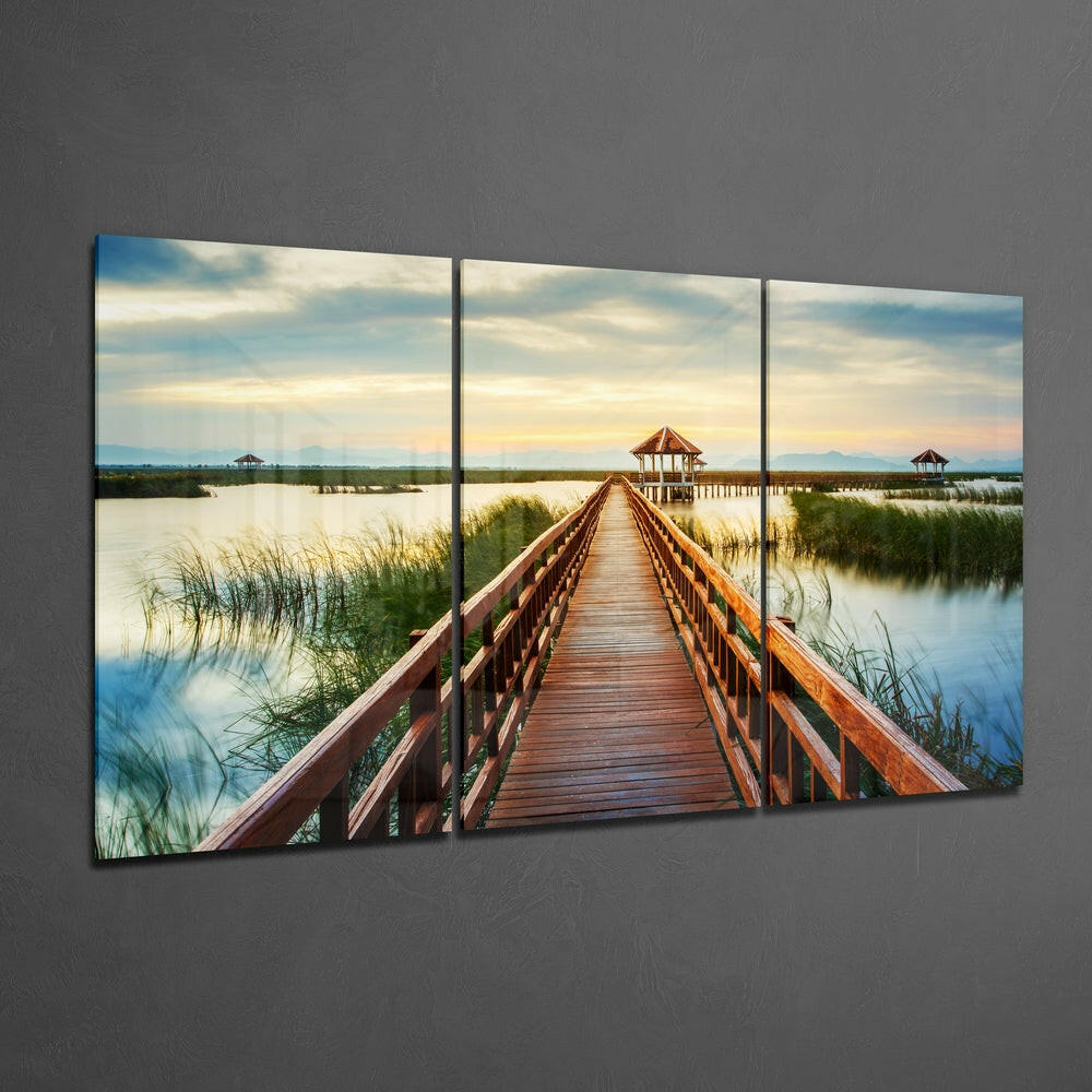 Three-panel glass wall art displaying a wooden boardwalk extending toward a gazebo across calm wetlands at sunset.