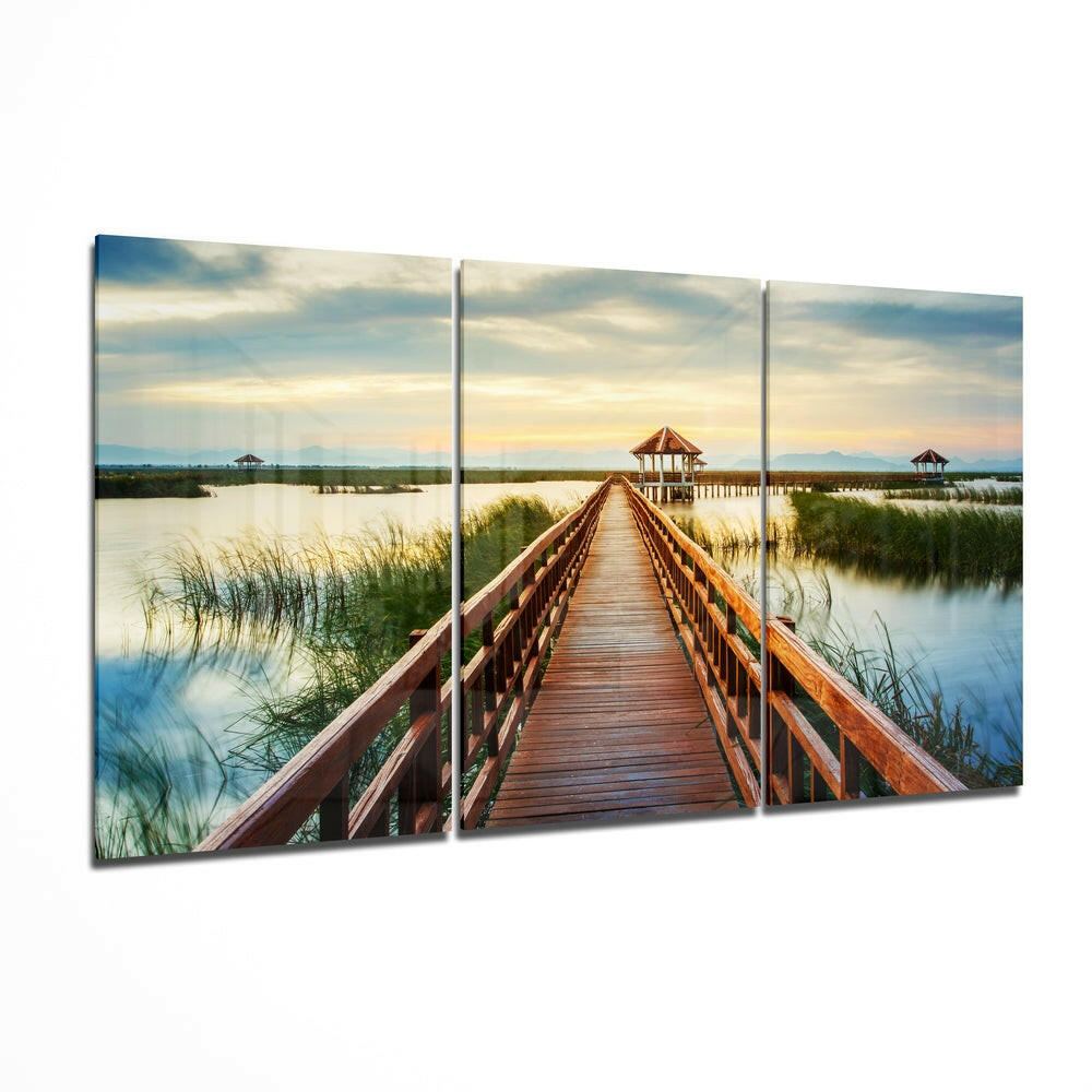 Three-panel glass wall art displaying a wooden boardwalk extending toward a gazebo over marshland and water at sunset.