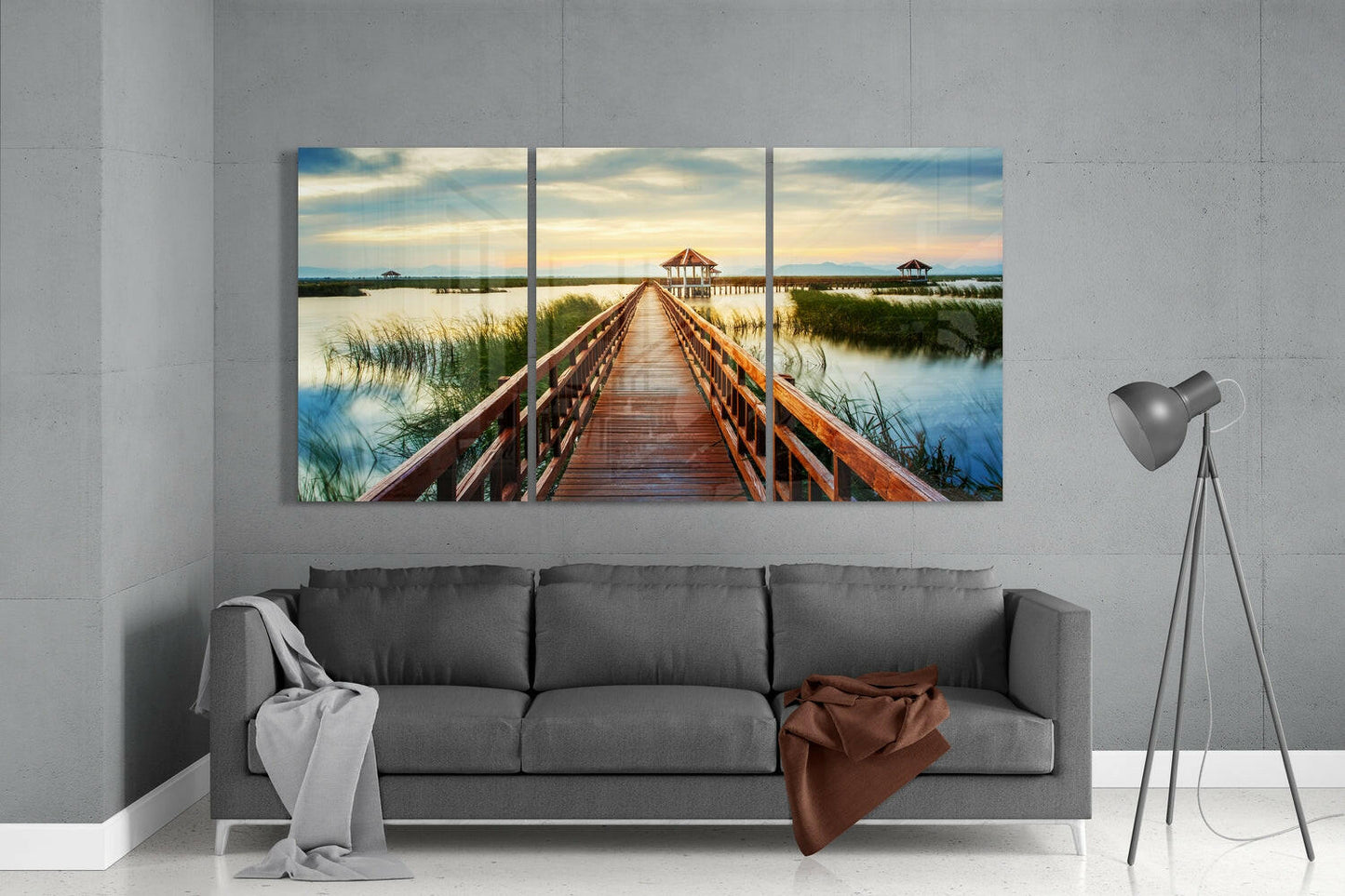 Three-panel glass wall art depicting a wooden boardwalk stretching toward gazebos across wetlands at sunset, displayed above a gray sofa.