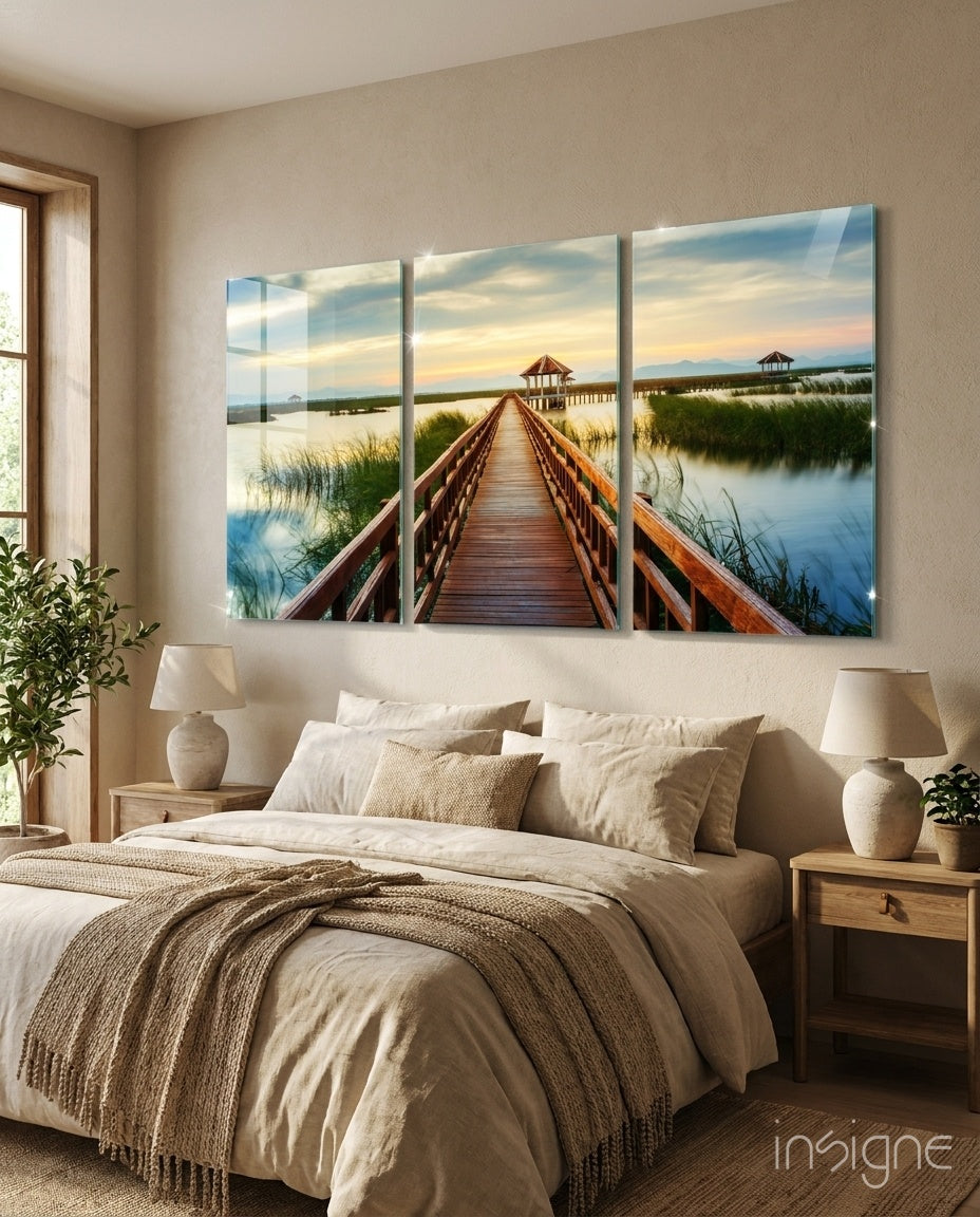 Three-panel glass wall art displaying a wooden boardwalk bridge extending toward a gazebo over marshland at sunset.