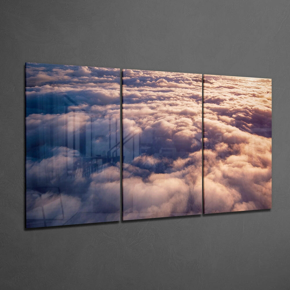 Three-panel glass wall art displaying an aerial view of clouds illuminated by golden sunlight, mounted on a gray textured wall.