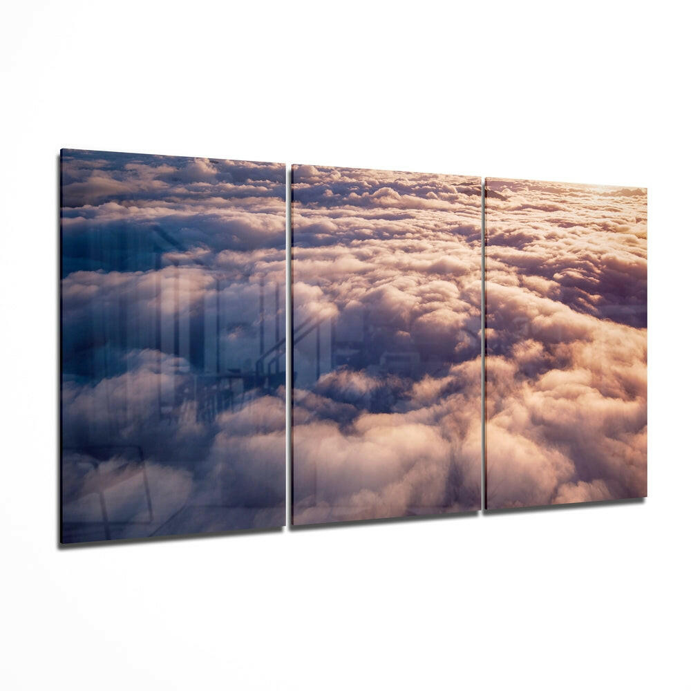 Three-panel glass wall art displaying an aerial view of golden-lit clouds with a city skyline visible through the mist on the left panel.