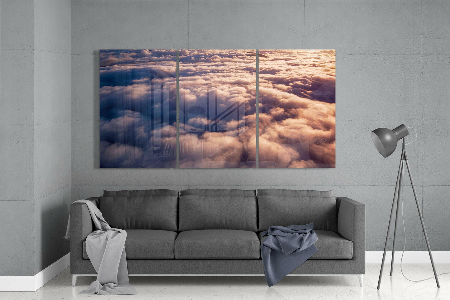 Three-panel glass wall art displaying golden-hour clouds above a cityscape, installed above a gray sofa in a minimalist interior.