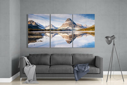 Three-panel glass wall art displaying mountain lake reflection with snow-capped peaks, displayed above gray sofa in modern living room.