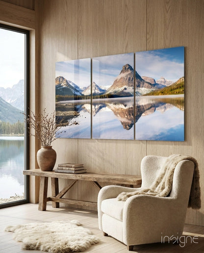 Three-panel glass wall art displaying snow-capped mountains and calm lake reflection in natural wood interior with cream chair.