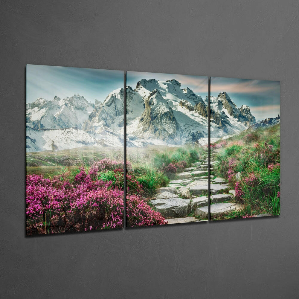 Three-panel glass wall art displaying a mountain landscape with snow-capped peaks, stone pathway, and vibrant pink wildflowers in foreground.