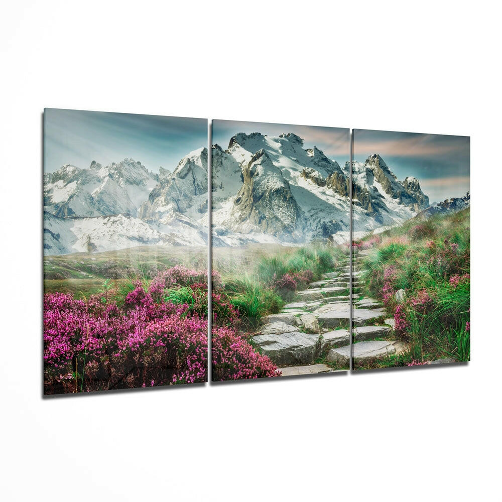 Three-panel glass wall art depicting a mountain landscape with snow-capped peaks, pink wildflowers, green grasses, and a stone pathway.
