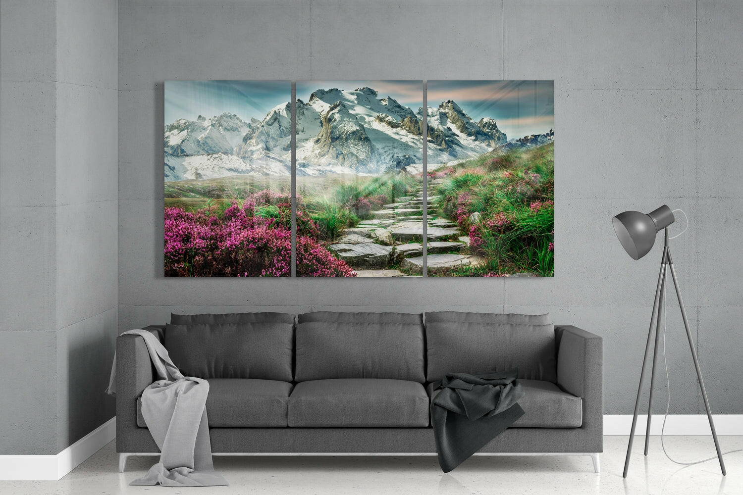 Three-panel glass wall art displaying snow-capped mountains and magenta wildflowers flanking a stone pathway, installed above gray sofa in modern interior.