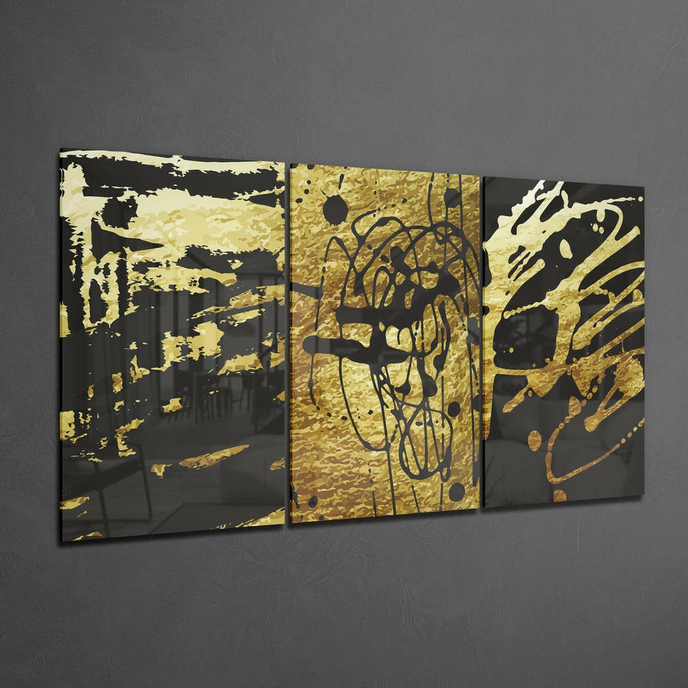 Three-panel black and gold abstract glass wall art with splatter and doodle designs displayed on gray wall.