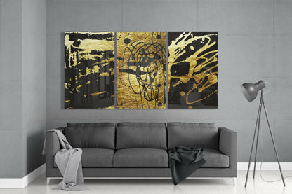 Three-panel abstract glass wall art with gold and black doodle designs displayed above gray sofa in modern living room.