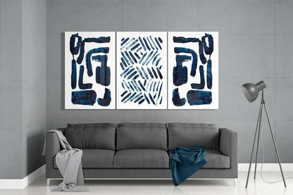 Three framed glass panels with navy blue abstract brushstrokes and geometric patterns hung above a gray sofa in a minimalist living room.