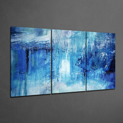 Three-panel abstract blue glass wall art mounted on gray wall, featuring vertical brushstroke patterns in deep and light blue tones.